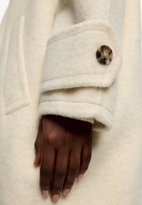 Manica di un cappotto di lana crema con un risvolto, caratterizzato da un bottone rotondo e decorato. Una mano appoggiata sul risvolto mostra una texture della pelle liscia.