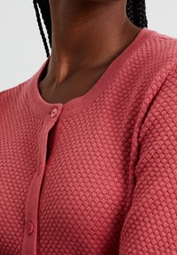 Růžový texturovaný kardigan s medovým vzorem, s kulatým výstřihem a zapínáním na knoflíky vpředu. Měkký pletený materiál, těsně přiléhající střih.