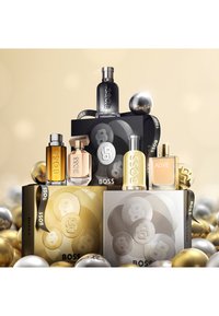 Parfumset tentoongesteld met vijf glazen flessen in diverse vormen en kleuren, omringd door zilveren en gouden decoratieve bollen. De dozen bevatten gedurfde logo's.