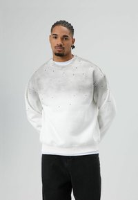 Witte sweatshirt met een gradiëntgrijze effect, voorzien van verspreide zwarte stippen, ronde halslijn en geribbelde manchetten. Draagbaar met zwarte broek.