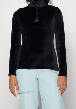 Femme portant un haut polaire noir à manches longues avec fermeture éclair et logo araignée sur la poitrine, et un pantalon bleu clair avec poche zippée.