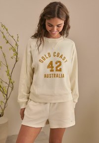 Svettr med krämfärgad nyans och texturerad tyg, med "GOLD COAST 42 AUSTRALIA" i guldbokstäver. Matchas med shorts i samma färg.