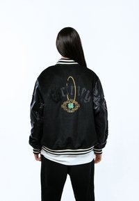 Chaqueta bomber negra con mangas de cuero, cuello blanco a rayas y diseño floral verde bordado con texto en la parte posterior.