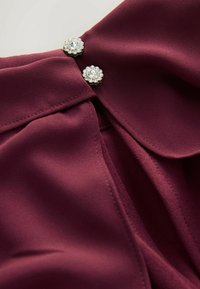 Bordeaux bluse i blødt materiale med to dekorative sølvknap detaljer og en foldet kravedesign.