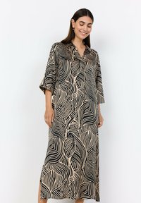 Lång svart klänning med beige bladmönster, knäppning fram, och trekvartsärmar. Mjukt, flödande tyg och sidofickor för lätt rörelse.