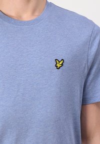Svetlomodré tričko s okrúhlym výstrihom a malým žltým vyšívaným logom vtáka na ľavej strane hrudníka.
