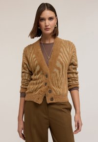 Nainen, jolla on päällä ruskea pörröinen nappikauluksellinen neuletakki, jossa on tekstuuri, pitkähihaisen paidan päällä, yhdistettynä ruskeisiin housuihin, seisomassa neutraalilla taustalla.