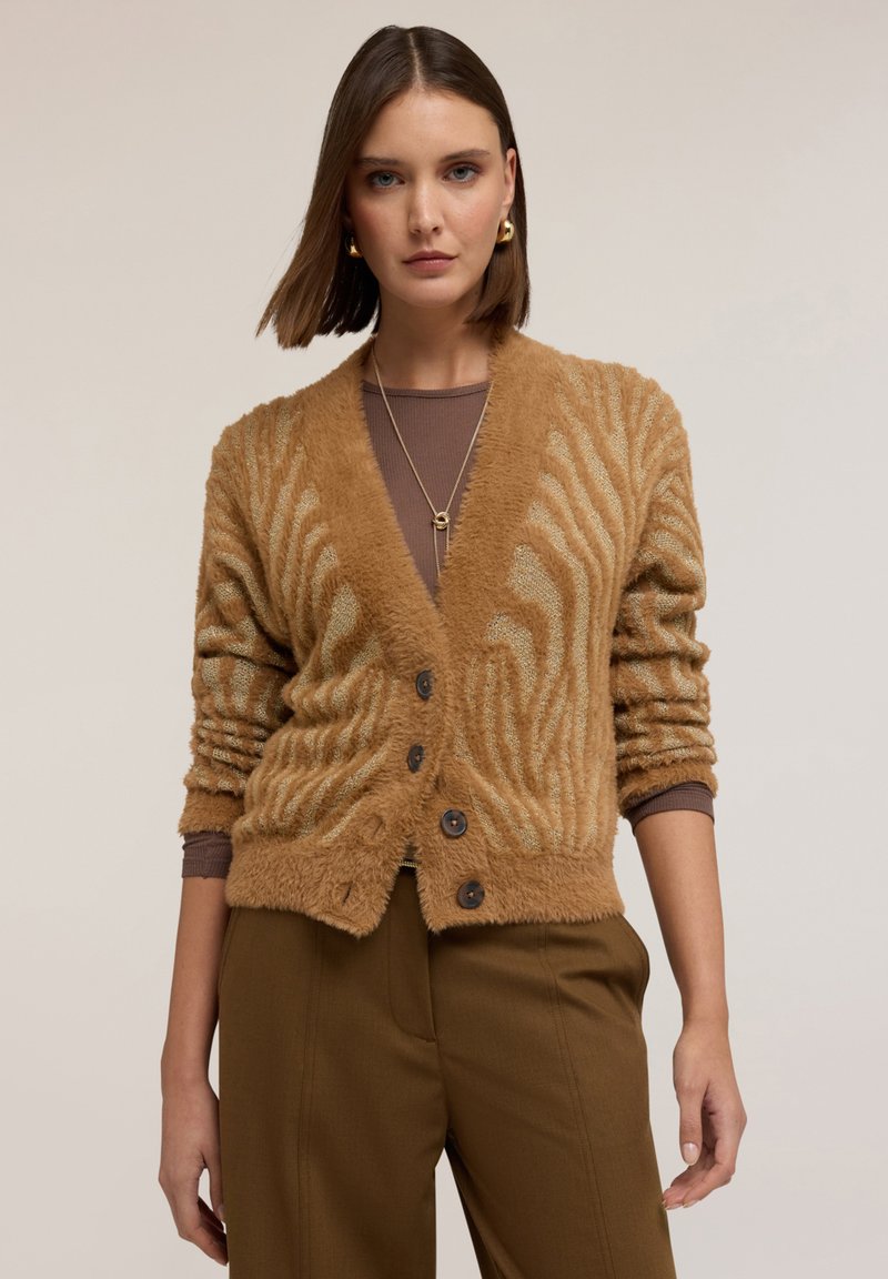 Nainen, jolla on päällä ruskea pörröinen nappikauluksellinen neuletakki, jossa on tekstuuri, pitkähihaisen paidan päällä, yhdistettynä ruskeisiin housuihin, seisomassa neutraalilla taustalla.