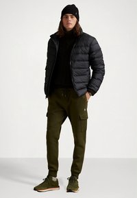 Czarna ocieplana kurtka, zielone spodnie bojówki, czarny sweter z dzianiny, czarna czapka, szare skarpetki i zielone trampki. Casualowy zimowy strój.