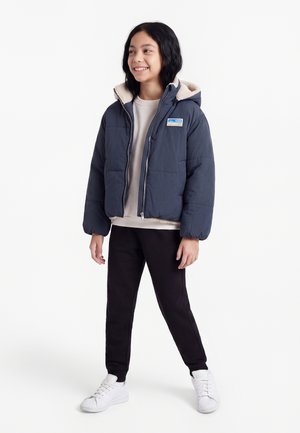 Smilende barn med skulderlangt sort hår iført en navyblå hættevest, beige skjorte, sorte bukser og hvide sneakers.