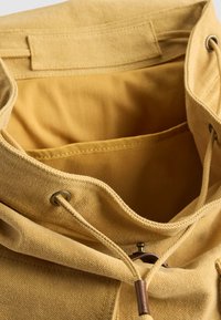 Beige canvas rugzak met open top, koordsluiting, metalen oogjes en een binnenvak zichtbaar in het hoofdcompartiment.