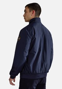 Navyblå jacka i lätt tyg med hög krage, elastiska ärmmuddar och ribbad midja. Har ett cirkulärt emblem på ärmen.
