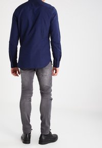 Hombre de pie, de espaldas, vistiendo una camisa de manga larga azul marino, jeans gris claros deslavados y zapatos de cuero negros, sobre un fondo blanco.