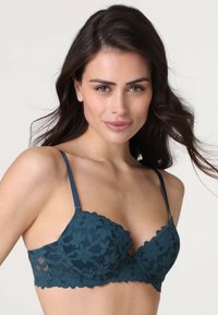 Reggiseno in pizzo color turchese con motivo floreale, coppe imbottite, bordi ondulati e spalline regolabili. Texture liscia con dettagli trasparenti sui lati.