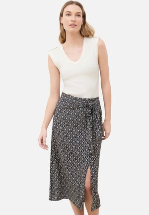 Jeune femme portant un haut blanc sans manches et une jupe midi noire à motifs avec une fente devant et un lien à la taille, debout devant un fond uni.