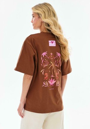 Blondynka o długich włosach, ubrana w brązowy t-shirt z różowymi tropikalnymi roślinami i wzorami tygrysa na plecach.