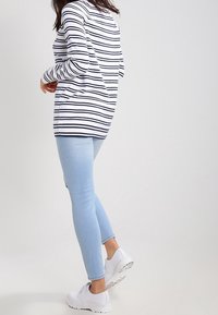 Persona che indossa un maglione a righe bianche e nere, jeans skinny blu chiaro e sneaker bianche, in piedi con un piede leggermente sollevato.
