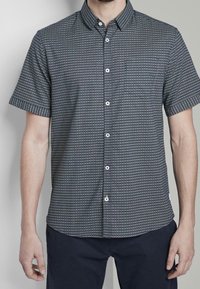 Uomo che indossa una camicia a maniche corte con bottoni, caratterizzata da un piccolo motivo geometrico in colori scuri e chiari, abbinata a pantaloni scuri.