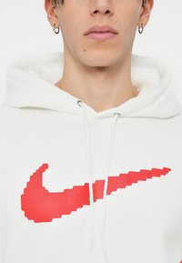 Bela kapuca z velikim, pikseliranim rdečim logotipom Nike swoosh. Kapuca z vrvicami in srebrnim okovjem. Gladek tekstilni videz.
