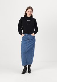 Hanorac negru cu sireturi și logo, purtat cu o fustă lungă din denim albastru. Modelul poartă cizme negre până la glezne, cu mâinile în buzunare. Look simplu, casual.