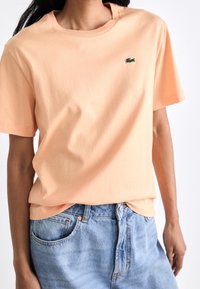 Ferskenfarvet bomuld T-shirt med rund halsudskæring, korte ærmer og en lille grøn logo på brystet, kombineret med lyseblå denimshorts.