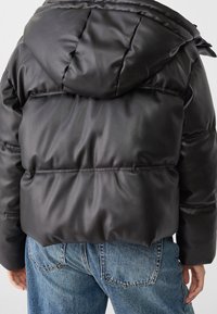 Black puffer jacket with a glossy finish, featuring a hood, quilted design, and cinched waist. Worn over light-wash denim jeans.