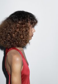 Červený saténový top s hlubokým výstřihem ve tvaru V, předvádějící hladkou texturu; modelka s objemnými kudrnatými vlasy pózuje na bílém pozadí.