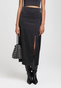 Jupe midi en satin noir avec une fente à l'avant, associée à un petit sac à carreaux et des bottes noires haute genoux. Texture lisse, design élégant.
