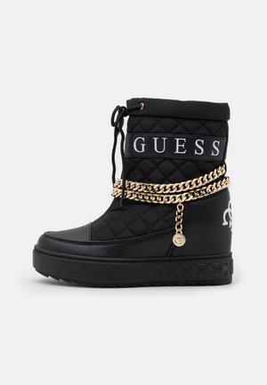 Botte d'hiver noire matelassée avec le logo "GUESS", chaîne dorée autour de la cheville et semelle en caoutchouc à semelle texturée.