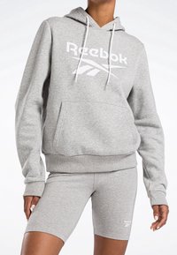 Felpa grigia Reebok con logo bianco, in materiale felpato, con cappuccio regolabile e tasca a marsupio. Abbinata a pantaloni corti grigi coordinati con dettaglio del logo.