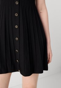 Jupe plissée noire avec nervures verticales et boutons dorés décoratifs, montrée sur le bas du torse et le bras d'une personne sur un fond uni.