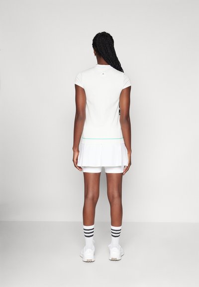 Fehér sport felső rövid ujjakkal és pliszírozott szoknyával. Finom zöld csíkkal díszített, fehér rövidnadrággal és csíkos zoknival viselve.