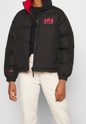 Modelo con una chaqueta acolchada negra de Helly Hansen, con forro rojo y logotipo, combinada con un top negro y unos pantalones blancos.