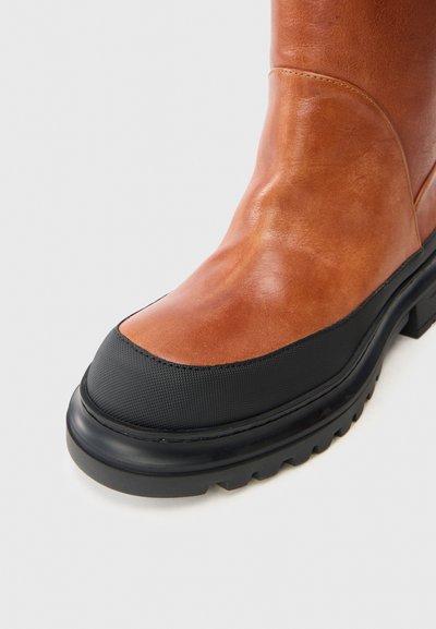 Καφέ δερμάτινη μπότα μέχρι τον αστράγαλο με μαύρη καπάκι από καουτσούκ στο πόδι και υφασματώδεις λεπτομέρειες, διαθέτοντας χοντρή μαύρη σόλα και λεία φινίρισμα στο δέρμα.