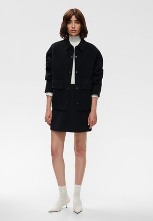 Femme portant une veste noire boutonnée, une jupe noire, un haut col roulé blanc, des chaussettes blanches et des chaussures blanches à talons pointus, debout sur fond gris.