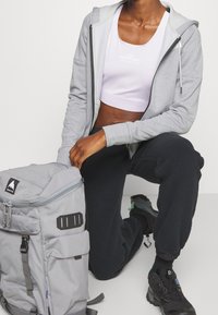 Persona de rodillas vestida con una sudadera gris con cremallera, una camiseta blanca corta, pantalones de forro polar negros, zapatillas negras, sosteniendo una mochila grande gris.