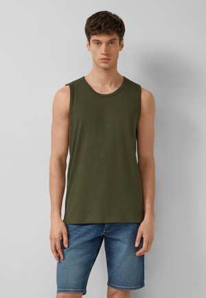 Jonge man met een olijfgroen mouwloos shirt en blauwe denim shorts, staand tegen een effen grijze achtergrond.