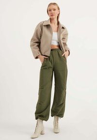 Beige cropped jas met een kraag, gecombineerd met losse groene broeken en crème veterlaarzen met hak. Glad materiaal met een relaxte pasvorm.