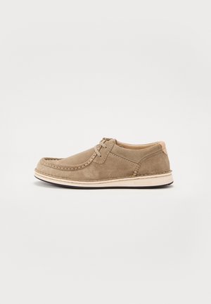 Scarpa casual beige in camoscio con allacciatura, design moc toe e suola color crema su sfondo bianco.