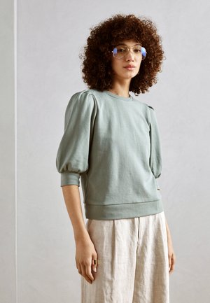 Hellgrüner Sweatshirt mit puffärmel, rundem Halsausschnitt und geripptem Saum. Kombiniert mit beigen, locker sitzenden Hosen. Glatte Textur.