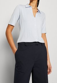Donna che indossa una camicia blu chiaro a maniche corte, infilata in pantaloni neri a vita alta, con le mani nelle tasche.