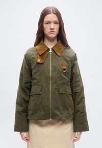 Young woman wearing an olive green jacket with brown corduroy collar and gold zipper, standing against a plain light background.