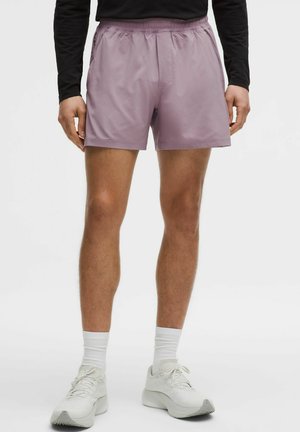 Man draagt mauve sportshorts, witte sokken en witte sneakers, staat met handen ontspannen langs zijn zij.