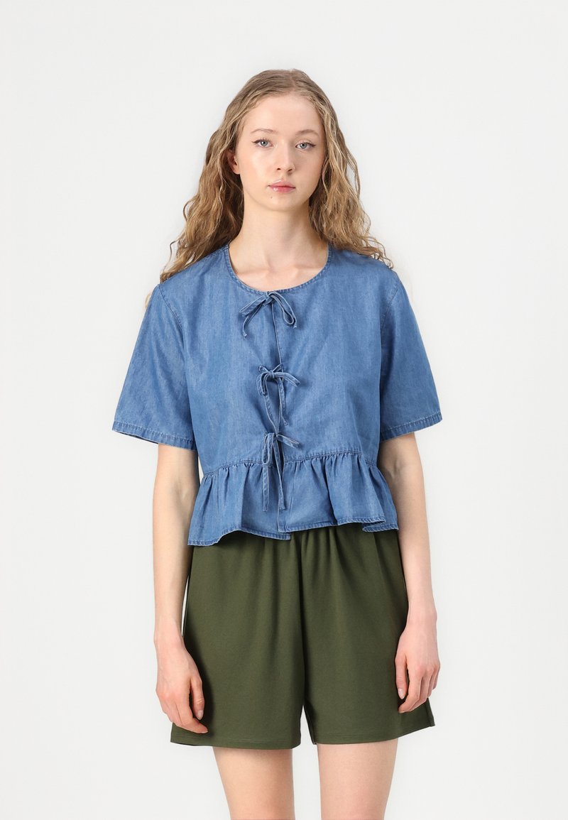 Svetlo modra denim srajca s kratkimi rokavi, peplum rob in sprednje vezanje; v kombinaciji s temno zelenimi, ohlapnimi kratkimi hlačami. Preprost dizajn.