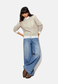 Pull en maille beige avec un motif texturé, superposé à une chemise blanche à bordure en dentelle. Jean en denim à jambes larges et chaussures à imprimé léopard.