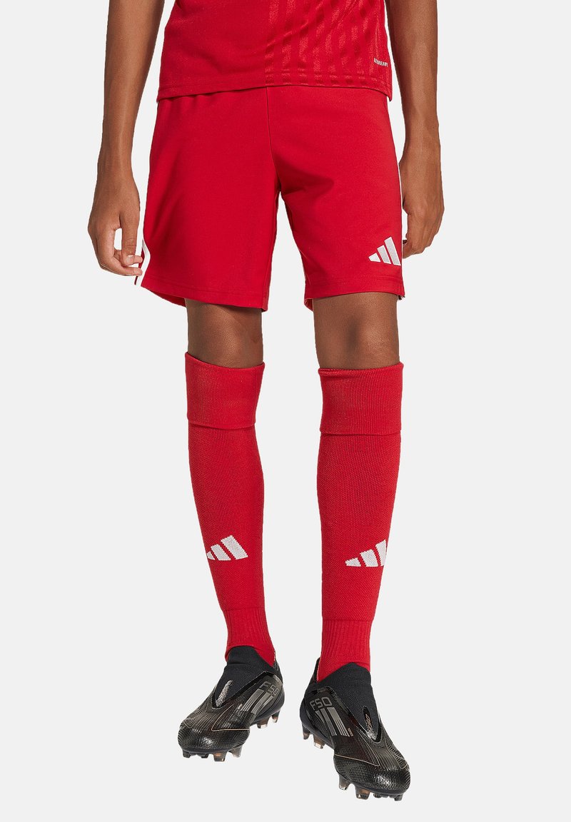 Rode voetbal shorts gemaakt van ademend materiaal, gecombineerd met rode kniekousen met witte logo-accenten, en zwarte voetbalschoenen.