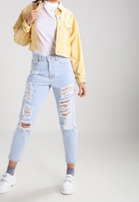 Veste cropped jaune, t-shirt blanc, jean clair déchiré avec de grandes déchirures, porté avec des baskets blanches et des chaussettes grises.