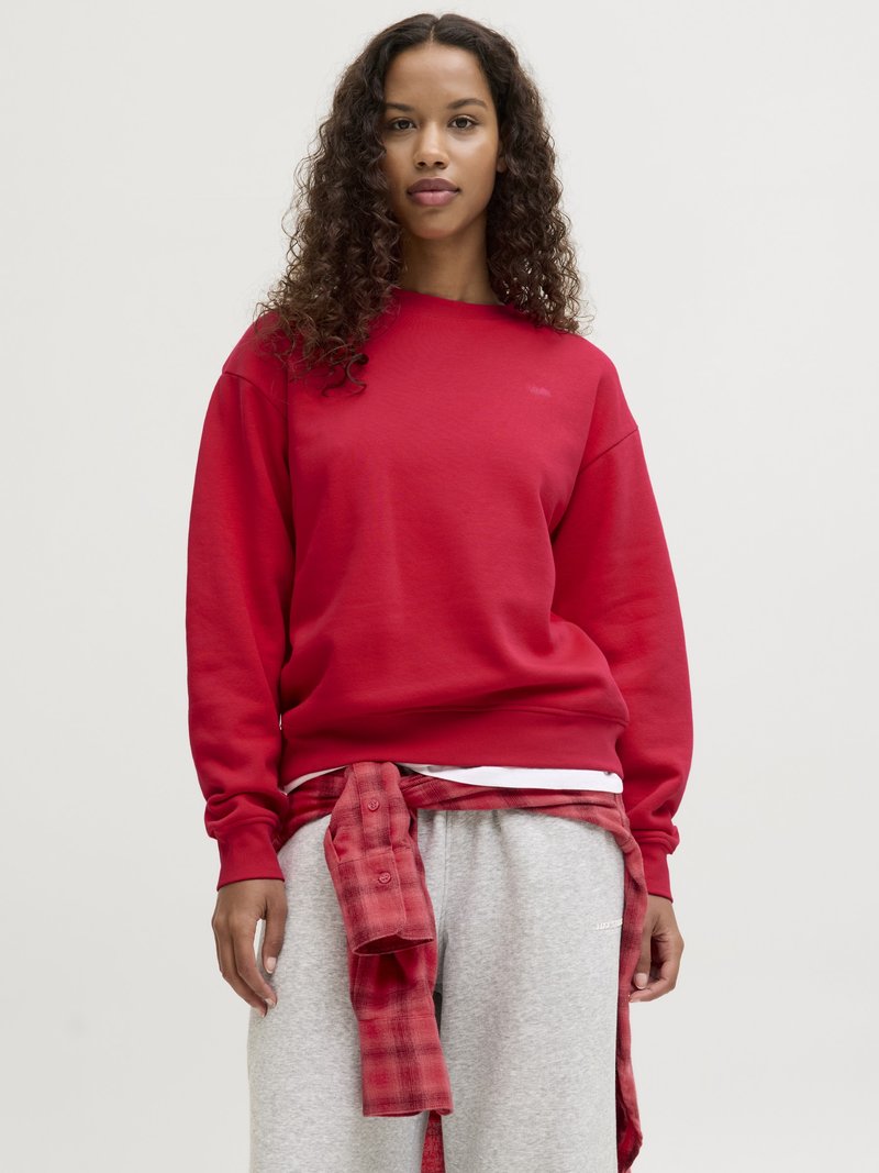 Kvinne med krøllete hår iført en rød genser, grå joggebukser og en rød rutete skjorte knytt rundt livet, stående foran en ensfarget bakgrunn.