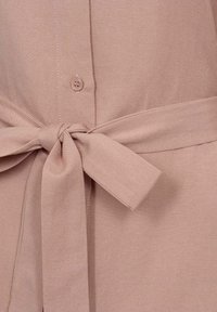 Camisa de tejido beige con cierre frontal de botones y un cinturón atado en la cintura. La textura es suave y con un ligero brillo.