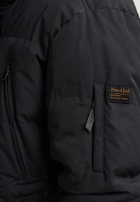 Zwarte pufferjack met een quilted textuur. Heeft een zichtbare ritssluiting en een logopatch op de mouw. Het materiaal lijkt waterafstotend.